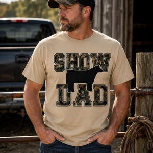 Può includere: Una t-shirt beige a maniche corte con il testo SHOW DAD in un carattere a blocchi con motivo mimetico. Una sagoma nera solida di una mucca è posizionata tra le parole SHOW e DAD sulla parte anteriore del capo.