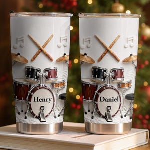 May include: Two white stainless steel tumblers featuring a drum set graphic with crossed drumsticks and musical notes. The bass drum on the left tumbler is personalised with the name Henry, and the bass drum on the right tumbler says Daniel.