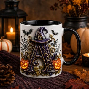 Pode incluir: Uma caneca de cerâmica branca com interior e alça pretos, apresentando uma letra A roxa grande e em relevo, adornada com um chapéu de bruxa, teias de aranha, morcegos e duas abóboras laranja brilhantes com rostos esculpidos.