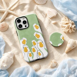 Puede incluir: Una funda de smartphone color verde salvia y un soporte circular a juego con un patrón repetitivo de patos blancos con picos amarillos. La funda tiene un recorte negro para la cámara y el diseño cubre toda la superficie posterior.