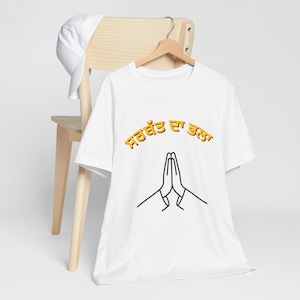 Puede incluir: Una camiseta blanca de manga corta con un gráfico de manos juntas en posición de oración y el texto en punjabi naranja "ਸਰਬੱਤ ਦਾ ਭਲਾ" arqueado encima. Una gorra de béisbol blanca lisa está colocada sobre la parte superior de la silla de madera.