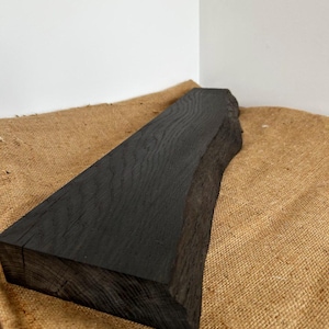 May include: A rectangular piece of dark stained wood with a natural live edge on one side. The wood features a visible grain pattern throughout its surface, showcasing a deep black finish that highlights the texture of the timber material.