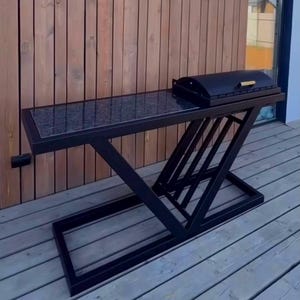 Peut inclure: Une station de barbecue d'extérieur en métal noir dotée d'un cadre rectangulaire avec une structure de support inclinée, un plan de travail en pierre tachetée gris foncé et un barbecue noir avec couvercle et poignée dorée.