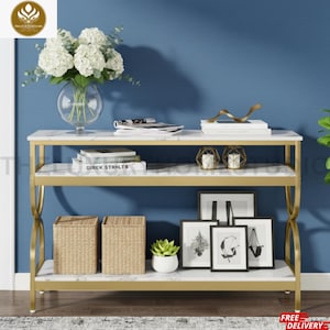 May include: A three-tier console table with a gold metal frame and white marble-patterned shelves. The table holds a glass vase with white flowers, books, decorative geometric candle holders, two woven storage baskets, and framed art prints.