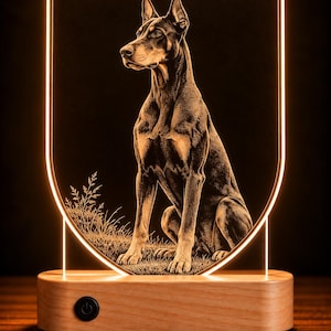 May include: An LED lamp featuring a shield-shaped acrylic panel with an etched illustration of a Doberman Pinscher sitting in grass. The panel is illuminated by a warm light and is mounted on a rectangular light-colored wooden base with a black power button.