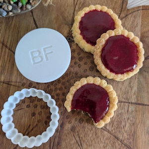 Op de afbeelding: Een witte ronde koekjesstempel met de letters BF in reliëf, een witte ronde koekjesvorm met geschulpte rand en drie ronde koekjes met geschulpte randen bedekt met donkerrode vruchtenjam, waarvan er één een hapje uit heeft.