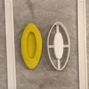 Peut inclure: Une découpe de pâte ou d'argile jaune en forme d'ovale à côté d'un emporte-pièce ovale en plastique blanc avec une barre centrale. Deux longues et fines bandes de plastique blanc sont placées verticalement de chaque côté des objets.