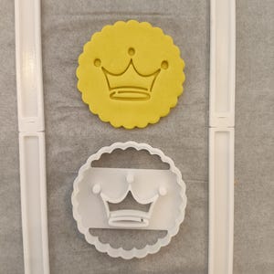 Peut inclure: Un biscuit circulaire jaune à bord festonné avec un motif de couronne en relief, placé au-dessus d'un emporte-pièce en plastique blanc présentant la même forme circulaire festonnée et le contour d'une couronne, entre deux rails de guidage.