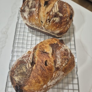 May include: Two oval-shaped loaves of artisan sourdough bread with deeply browned, flour-dusted crusts and distinct scoring marks. The loaves are resting on a rectangular silver-colored metal wire cooling rack with a grid pattern.