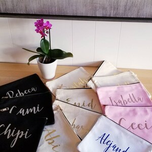 May include: A collection of personalized zippered pouches in various colors and fabrics. The pouches are decorated with names in gold, silver, and blue lettering. The names include "Rebecca", "Cami", "Chipi", "Abi", "Estaban", "Enid", "Harri", "Isabella", "Ceci", and "Alejandra".