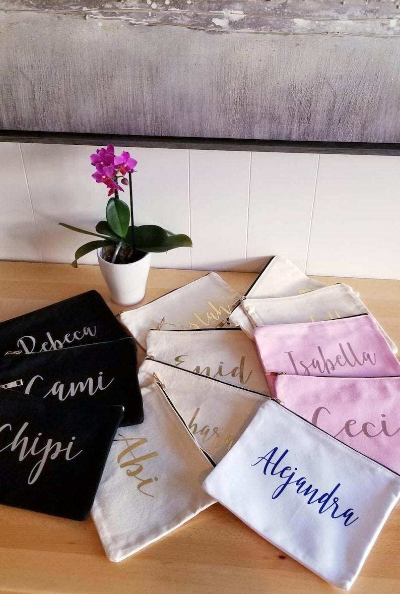 May include: A collection of personalized zippered pouches in various colors and fabrics. The pouches are decorated with names in gold, silver, and blue lettering. The names include "Rebecca", "Cami", "Chipi", "Abi", "Estaban", "Enid", "Harri", "Isabella", "Ceci", and "Alejandra".
