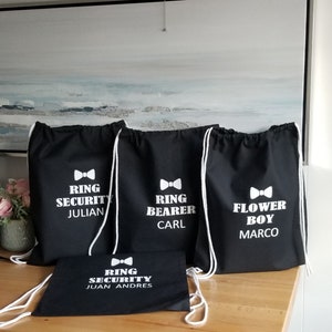 May include: Four black drawstring bags with white text and bowtie designs. The bags read "Ring Security Julian", "Ring Bearer Carl", "Flower Boy Marco", and "Ring Security Juan Andres".