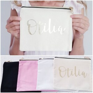 May include: Four zippered pouches in black, pink, white, and beige. The beige pouch has the name "Otilia" written in gold lettering.