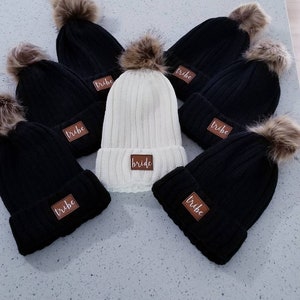 Puede incluir: Seis gorros de punto con pompones de piel sint&eacute;tica. Cinco gorros son negros con la palabra "tribe" en un parche marr&oacute;n. Un gorro es blanco con la palabra "bride" en un parche marr&oacute;n.