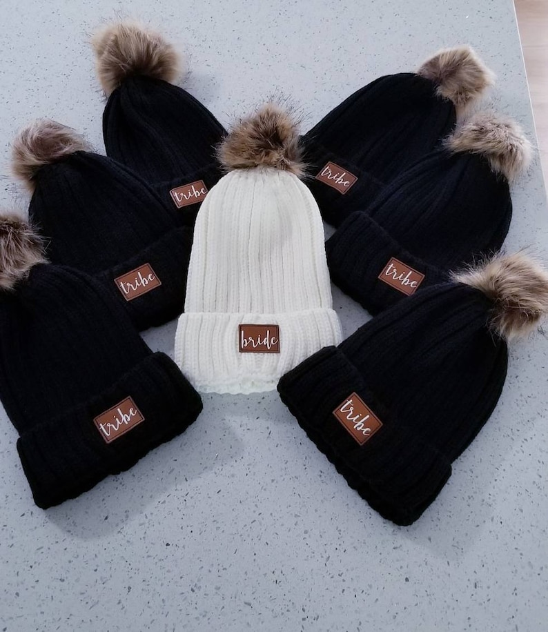 Puede incluir: Seis gorros de punto con pompones de piel sint&eacute;tica. Cinco gorros son negros con la palabra "tribe" en un parche marr&oacute;n. Un gorro es blanco con la palabra "bride" en un parche marr&oacute;n.