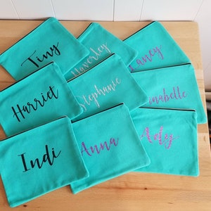 May include: A collection of teal zippered pouches with various names written in black and purple glitter on them. The names include: Tiny, Harriet, Indi, Anna, Adely,  Haverley, Stephanie,  Laney, and Isabelle.