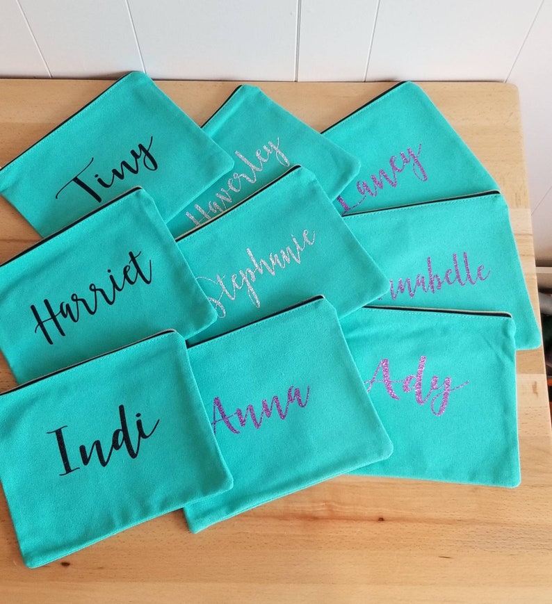 May include: A collection of teal zippered pouches with various names written in black and purple glitter on them. The names include: Tiny, Harriet, Indi, Anna, Adely,  Haverley, Stephanie,  Laney, and Isabelle.