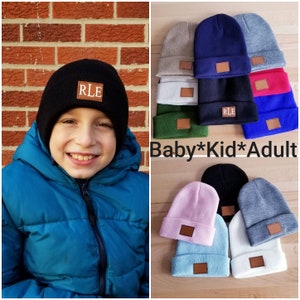 May include: A variety of knit beanies in different colors, including black, navy, gray, pink, blue, and white. Each beanie has a brown leather patch with a monogram on it. The text "Baby*Kid*Adult" is visible in the image.