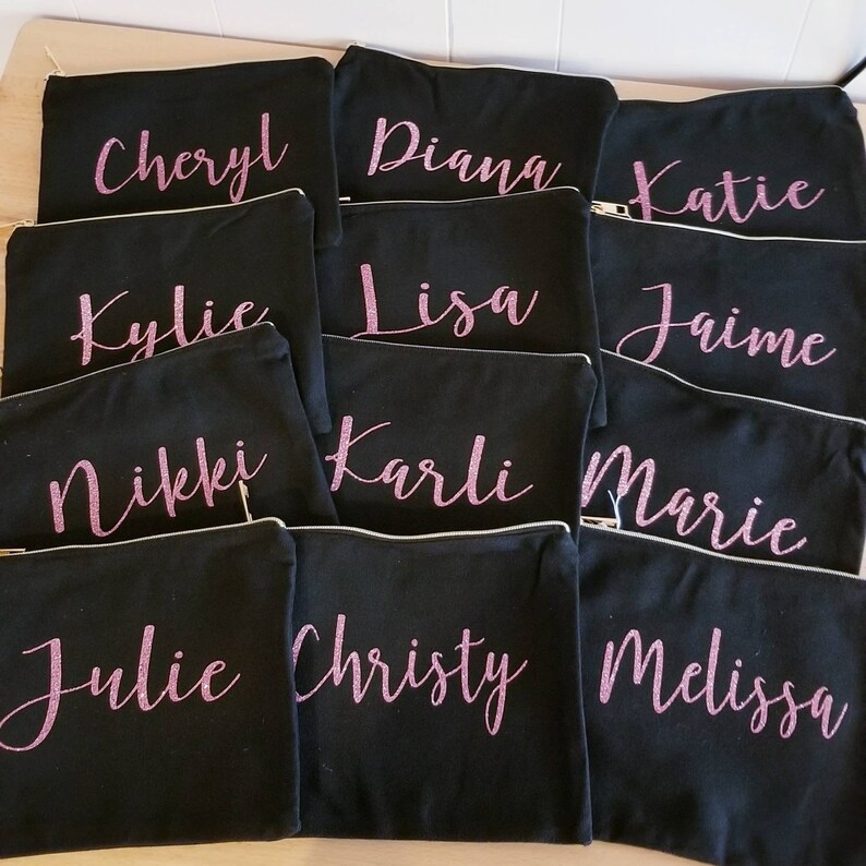 May include: Twelve black zippered pouches with pink glitter lettering. The pouches are arranged in three rows of four. The names Cheryl, Diana, Katie, Kylie, Lisa, Jaime, Nikki, Karli, Marie, Julie, Christy, and Melissa are written on the pouches.