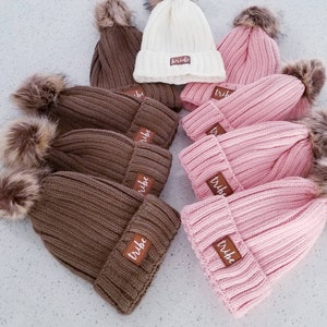 Puede incluir: Un grupo de gorros de punto marrones y rosas con pompones de piel sint&eacute;tica. Cada gorro tiene un parche de cuero marr&oacute;n con la palabra "tribe" escrita en blanco.