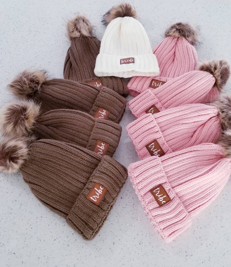 Puede incluir: Un grupo de gorros de punto marrones y rosas con pompones de piel sint&eacute;tica. Cada gorro tiene un parche de cuero marr&oacute;n con la palabra "tribe" escrita en blanco.