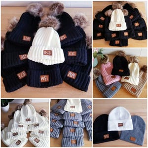 Puede incluir: Surtido de gorros de punto con pompones de piel sint&eacute;tica. Los gorros son de varios colores, incluyendo negro, blanco, gris, rosa y beige. Cada gorro tiene un parche de cuero con una letra o palabra diferente bordada, como "Bride", "KMT", "VL" y "MSL".