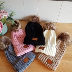 Puede incluir: Cinco gorros de invierno de punto con pompones de piel sint&eacute;tica. Los gorros son de varios colores, incluyendo rosa, negro, blanco, gris y marr&oacute;n. Cada gorro tiene un parche de cuero marr&oacute;n en el lateral.