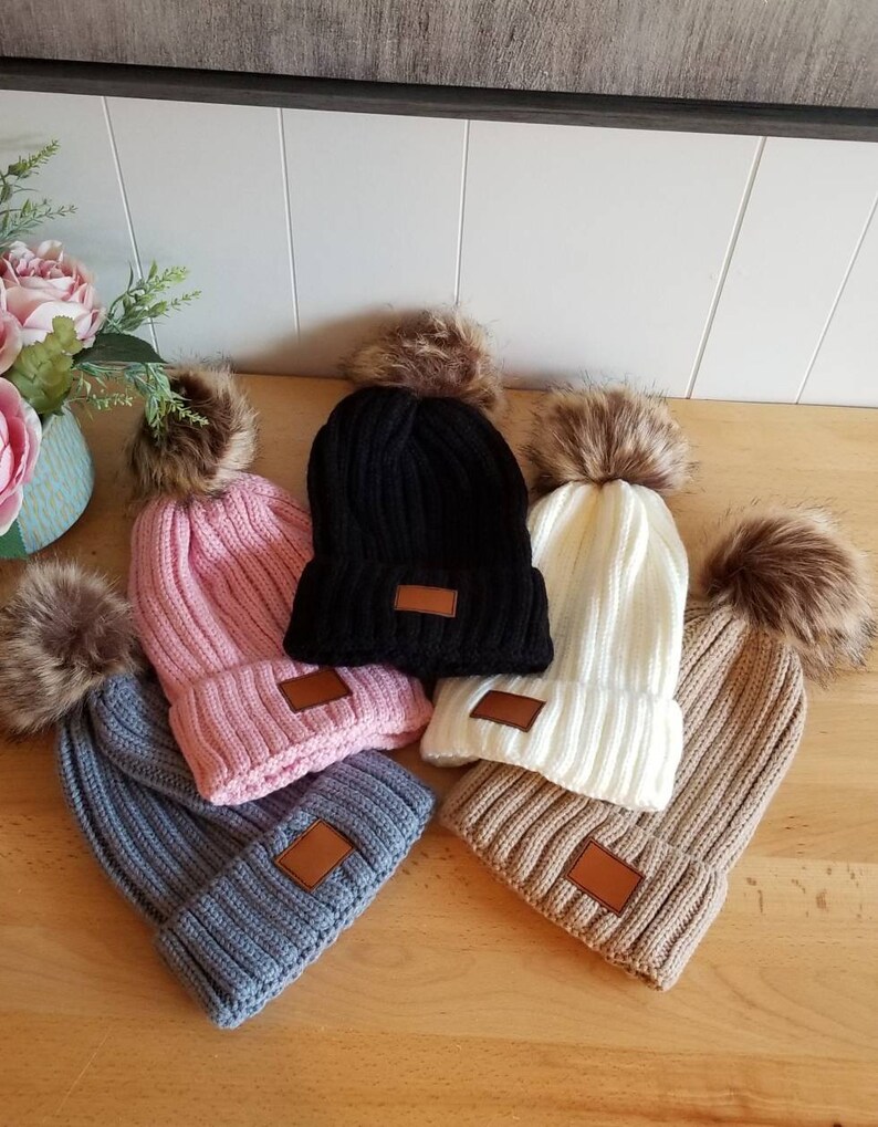 Puede incluir: Cinco gorros de invierno de punto con pompones de piel sint&eacute;tica. Los gorros son de varios colores, incluyendo rosa, negro, blanco, gris y marr&oacute;n. Cada gorro tiene un parche de cuero marr&oacute;n en el lateral.