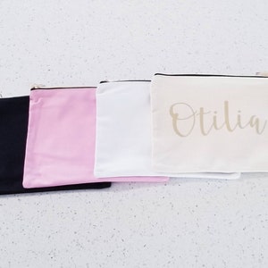 May include: Four zippered pouches in black, pink, white, and beige. The beige pouch has the name "Otilia" written in gold script.