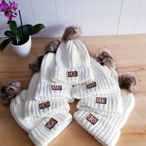 Puede incluir: Seis gorros de punto blanco con pompones de piel sint&eacute;tica marr&oacute;n. Cada gorro tiene un parche de cuero marr&oacute;n con iniciales bordadas en blanco. Las iniciales son CCC, KMT, CCH, AHY, HBC y CRD.