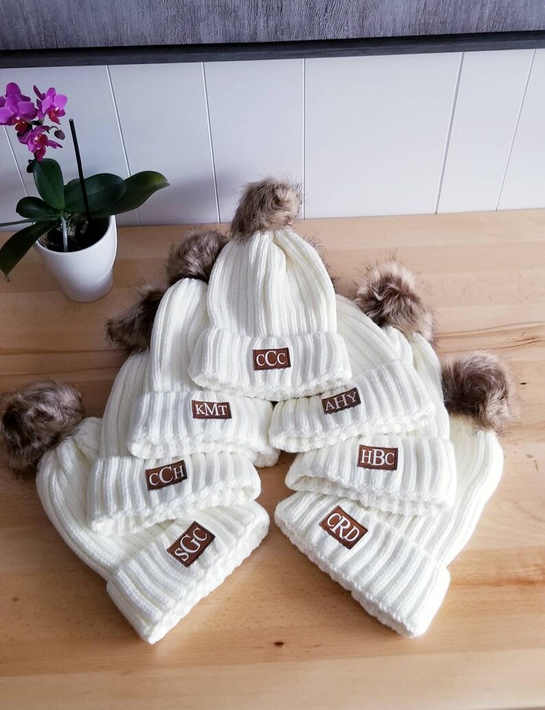 Puede incluir: Seis gorros de punto blanco con pompones de piel sint&eacute;tica marr&oacute;n. Cada gorro tiene un parche de cuero marr&oacute;n con iniciales bordadas en blanco. Las iniciales son CCC, KMT, CCH, AHY, HBC y CRD.
