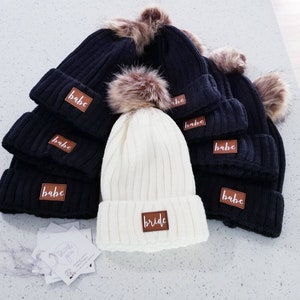 Puede incluir: Un conjunto de gorros de punto negros y blancos con pompones de piel sint&eacute;tica. Los gorros tienen parches de cuero marr&oacute;n con la palabra "babe" escrita en ellos. Un gorro es blanco y tiene un parche de cuero marr&oacute;n con la palabra "bride" escrita en &eacute;l.