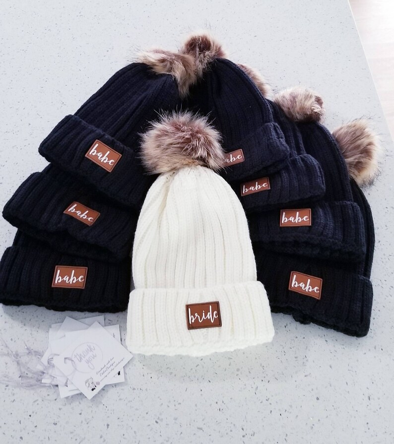 Puede incluir: Un conjunto de gorros de punto negros y blancos con pompones de piel sint&eacute;tica. Los gorros tienen parches de cuero marr&oacute;n con la palabra "babe" escrita en ellos. Un gorro es blanco y tiene un parche de cuero marr&oacute;n con la palabra "bride" escrita en &eacute;l.