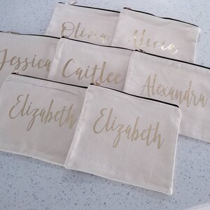 May include: Seven white canvas zippered pouches with gold lettering. The pouches are labeled with the names "Olivia", "Alicia", "Jessica", "Caitlee", "Alexandra", and "Elizabeth" (twice).