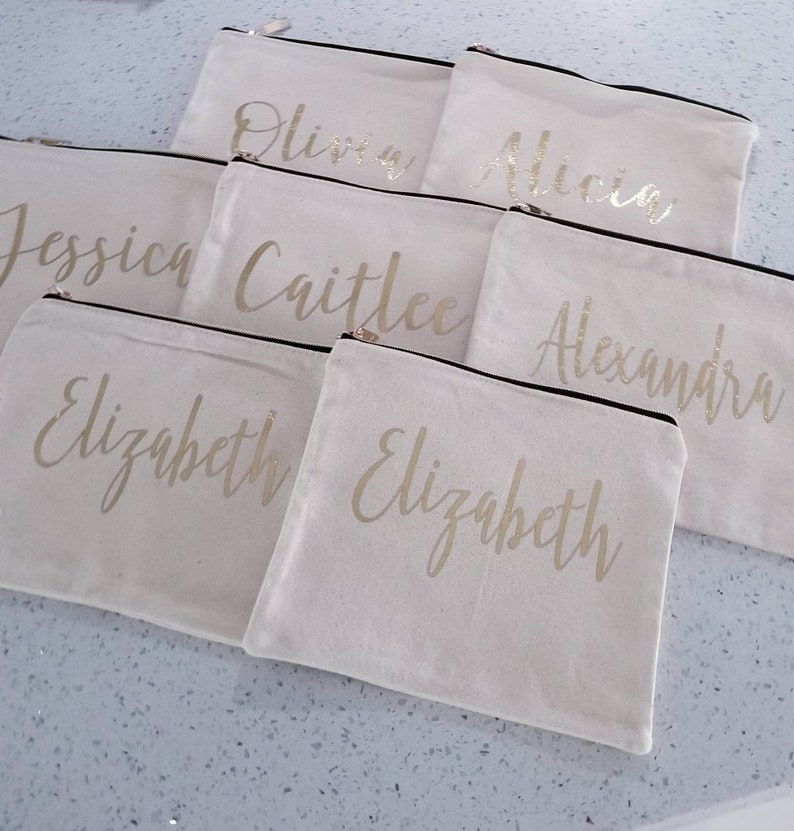 May include: Seven white canvas zippered pouches with gold lettering. The pouches are labeled with the names "Olivia", "Alicia", "Jessica", "Caitlee", "Alexandra", and "Elizabeth" (twice).