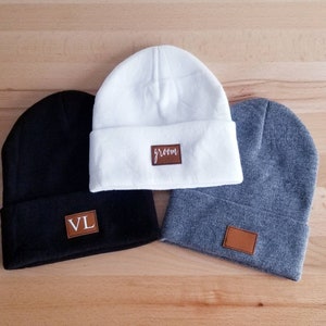 Puede incluir: Tres gorros de punto con parches de cuero. Un gorro es blanco con la palabra "groom" en el parche. Un gorro es negro con las iniciales "VL" en el parche. Un gorro es gris con un parche en blanco.