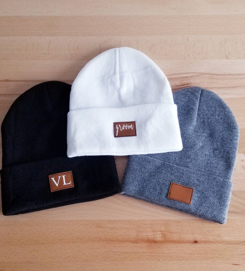 Puede incluir: Tres gorros de punto con parches de cuero. Un gorro es blanco con la palabra "groom" en el parche. Un gorro es negro con las iniciales "VL" en el parche. Un gorro es gris con un parche en blanco.