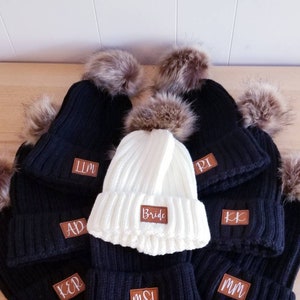 Puede incluir: Un conjunto de nueve gorros de punto negros y blancos con pompones de piel sint&eacute;tica. Los gorros tienen parches de cuero marr&oacute;n con letras blancas. Un gorro dice "Bride" y los dem&aacute;s tienen iniciales.