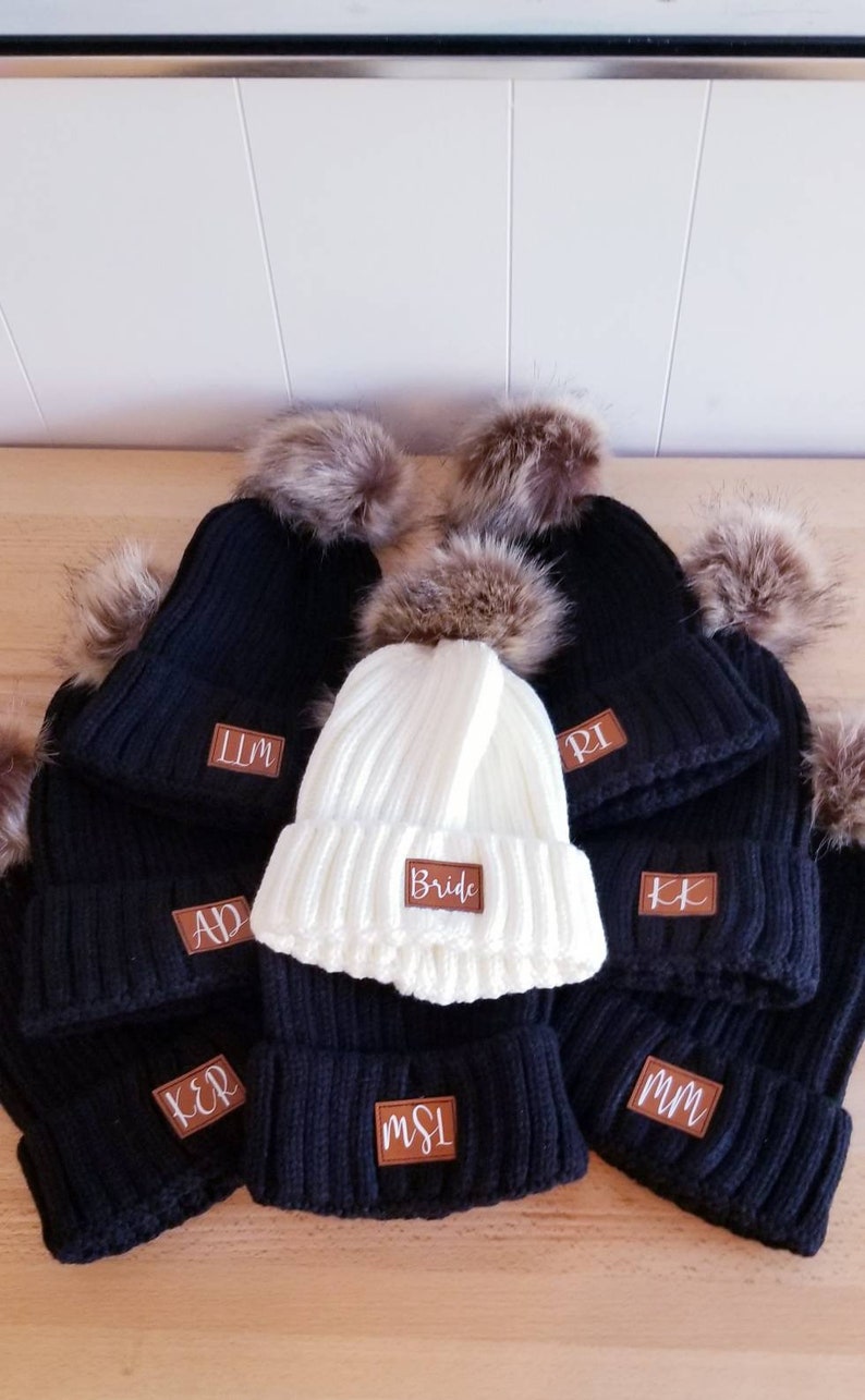 Puede incluir: Un conjunto de nueve gorros de punto negros y blancos con pompones de piel sint&eacute;tica. Los gorros tienen parches de cuero marr&oacute;n con letras blancas. Un gorro dice "Bride" y los dem&aacute;s tienen iniciales.