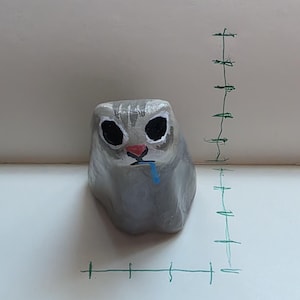 May include: A hand-painted gray rock shaped like a cat with large black eyes, a pink nose, and a blue tongue sticking out. The cat figure is positioned next to a hand-drawn green grid on the surface, showing a height of approximately 2.5 inches.