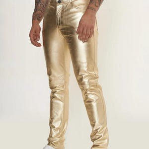 Könnte beinhalten: Metallic-goldene Hose mit schmaler Passform, einem klassischen Knopfverschluss an der Taille und sichtbaren horizontalen Nahtdetails an den Knien. Die Hose hat eine glatte, reflektierende Oberfläche und eine klassische Fünf-Taschen-Konstruktion.