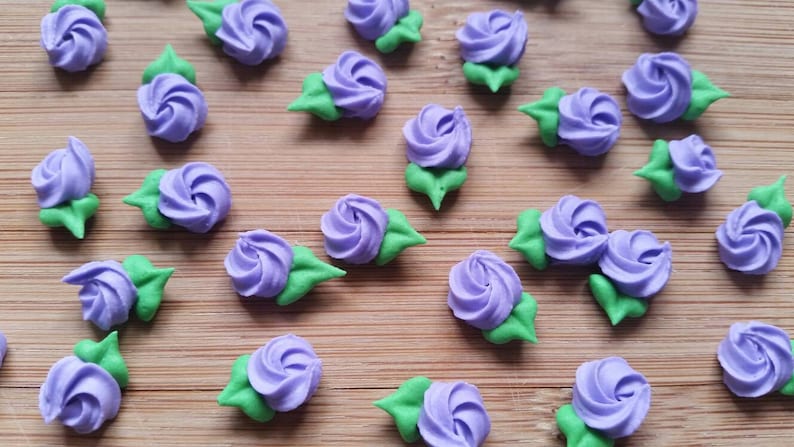 K&ouml;nnte beinhalten: Lila und gr&uuml;ne Zuckerblumen-Dekorationen. Die Blumen sind aus Zuckerguss und haben ein Spiraldesign. Sie sind auf einer Holzoberfl&auml;che angeordnet.