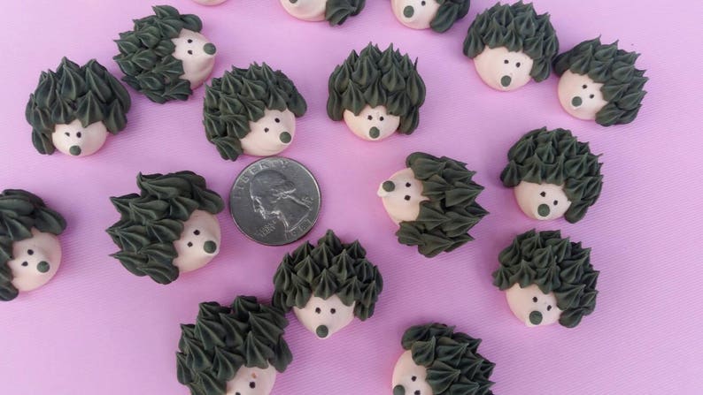 Puede incluir: Una colecci&oacute;n de peque&ntilde;os caramelos decorativos con forma de erizo, con espinas de glaseado verde oscuro y caras de color rosa claro. Se incluye una moneda para comparar el tama&ntilde;o, todo dispuesto sobre una superficie de color p&uacute;rpura claro.