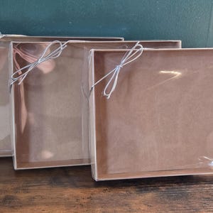May include: Three rectangular gift boxes with clear plastic lids are arranged on a wooden surface. Each box is brown with a silver wire bow. The boxes are empty and ready for use.