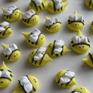 May include: A collection of yellow bee-shaped sugar cookies decorated with black stripes and white wings. The cookies are arranged on a white surface.