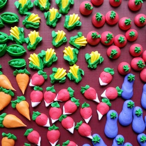 May include: Assortment of colorful buttercream frosting decorations shaped like vegetables, including peas in pods, corn on the cob, radishes, carrots, and eggplant.  The frosting decorations are arranged on a red surface.