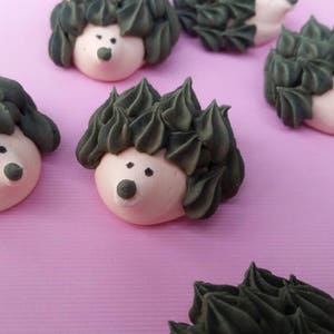 Puede incluir: Primer plano de varios pasteles con forma de erizo sobre una superficie rosa. Cada pastel tiene un cuerpo rosa claro, espinas de glaseado verde oscuro y peque&ntilde;os ojos y nariz de puntos negros. Los pasteles est&aacute;n dispuestos en un patr&oacute;n disperso.