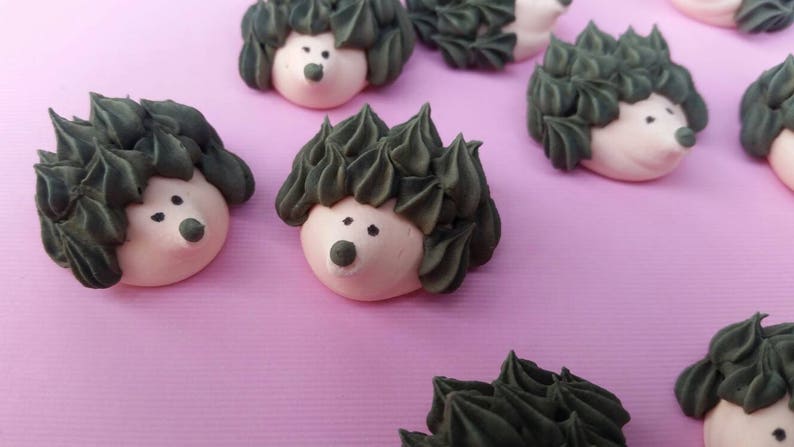 Puede incluir: Primer plano de varios pasteles con forma de erizo sobre una superficie rosa. Cada pastel tiene un cuerpo rosa claro, espinas de glaseado verde oscuro y peque&ntilde;os ojos y nariz de puntos negros. Los pasteles est&aacute;n dispuestos en un patr&oacute;n disperso.
