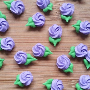 K&ouml;nnte beinhalten: Eine Sammlung von Zuckerblumen-Dekorationen in Lila und Gr&uuml;n. Die Blumen sind auf einer Holzoberfl&auml;che angeordnet.