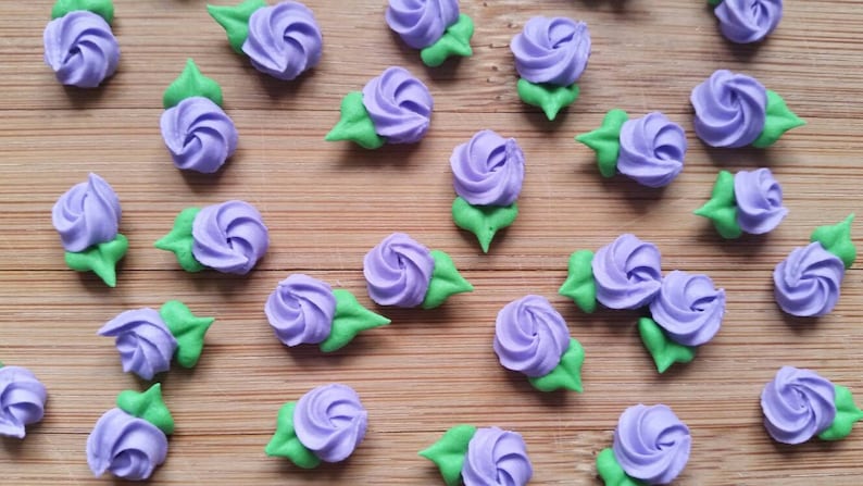 K&ouml;nnte beinhalten: Eine Sammlung von Zuckerblumen-Dekorationen in Lila und Gr&uuml;n. Die Blumen sind auf einer Holzoberfl&auml;che angeordnet.